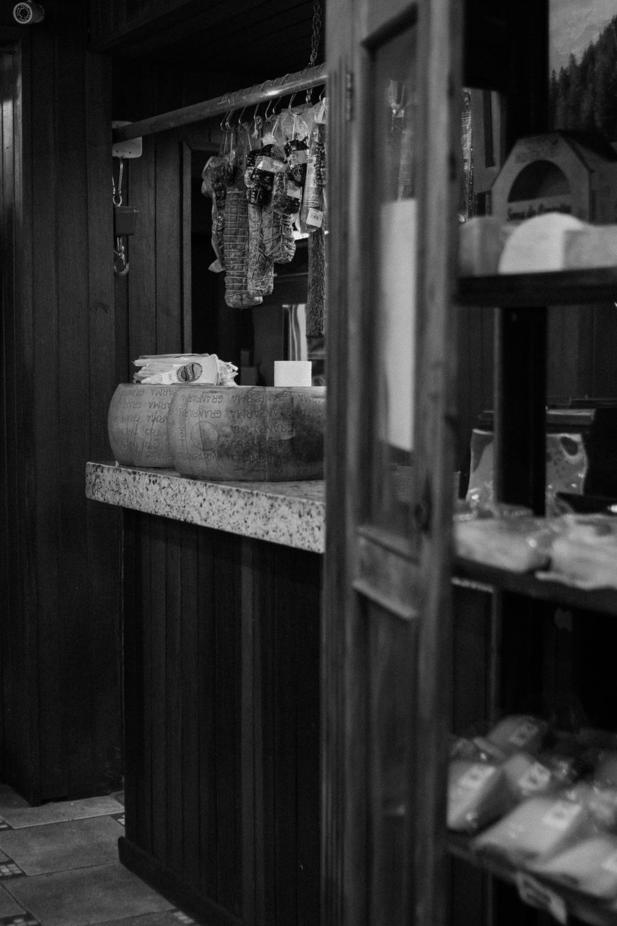 Black and white photo of cheese wheels and hanging cured meats in a rustic shop