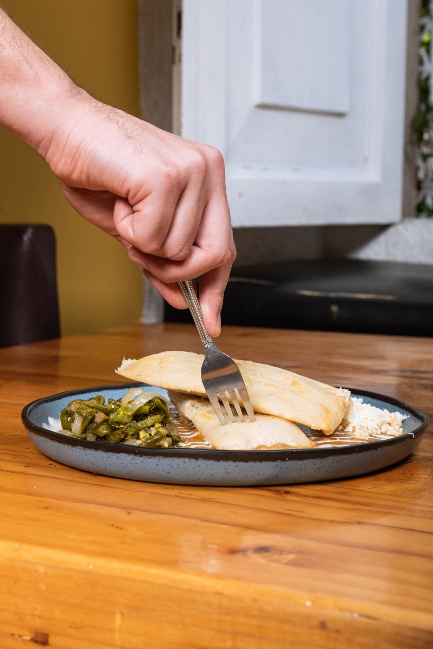 A hand holds a fork, cutting into a tamale served with vegetables