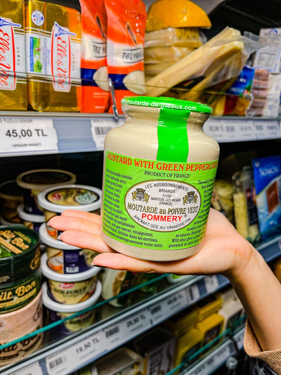 A hand holding a large jar of mustard in a grocery store aisle
