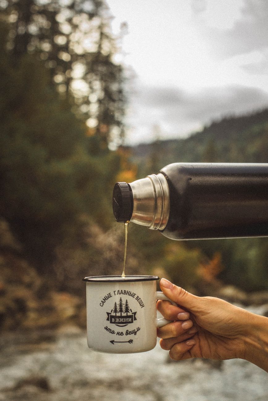 A hand pours coffee from a thermos into a mug in an outdoor forest setting