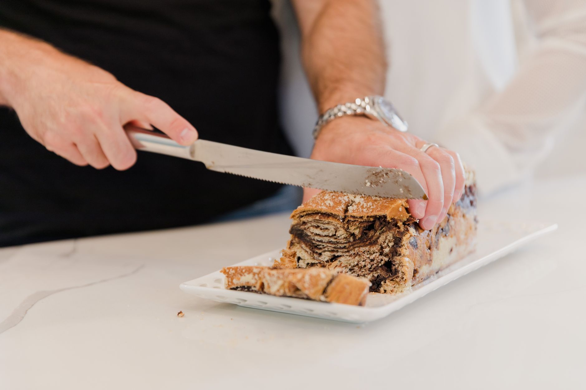 Close-up of hands slicing fresh chocolate babka bread on a white platter