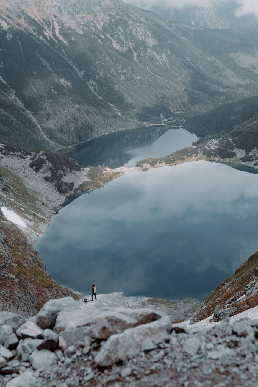 A lone hiker stands near a reflective mountain lake amidst scenic peaks