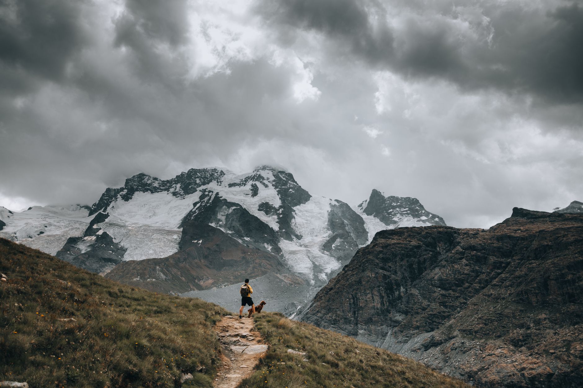 A man with his dog hikes along a trail with snow-covered mountains looming