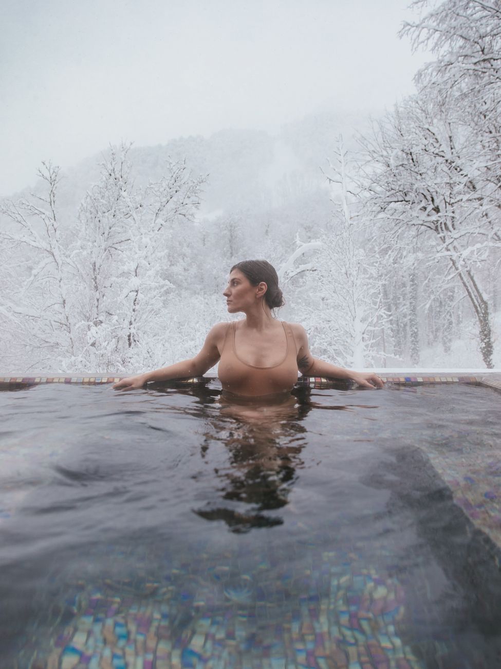 A woman enjoys a relaxing dip in an outdoor hot spring pool surrounded by snowy woods