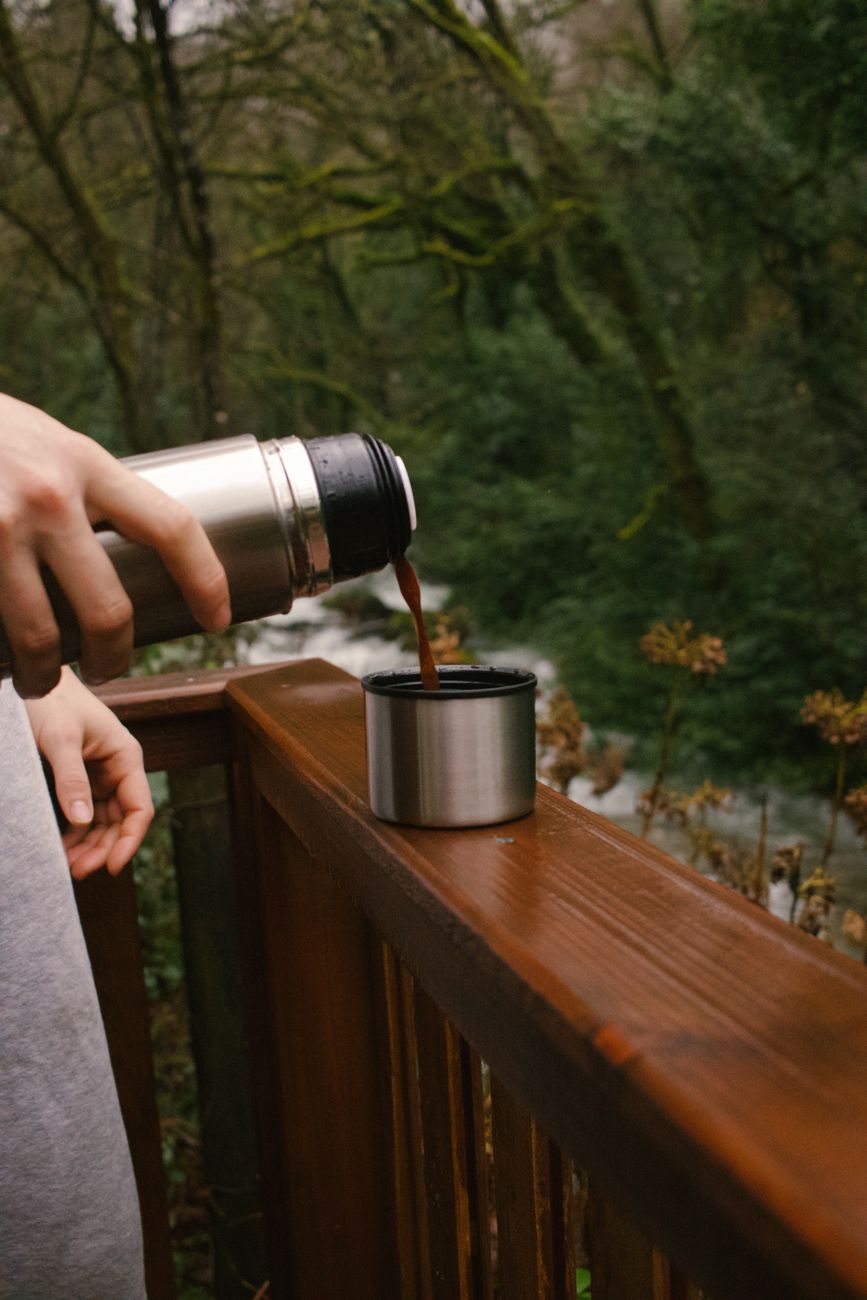 Person pouring coffee from a thermos in a serene outdoor wooded area